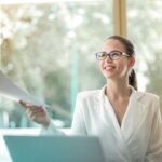 Low angle of successful female executive manager in classy style sitting at table with laptop in contemporary workplace and passing documents to colleague